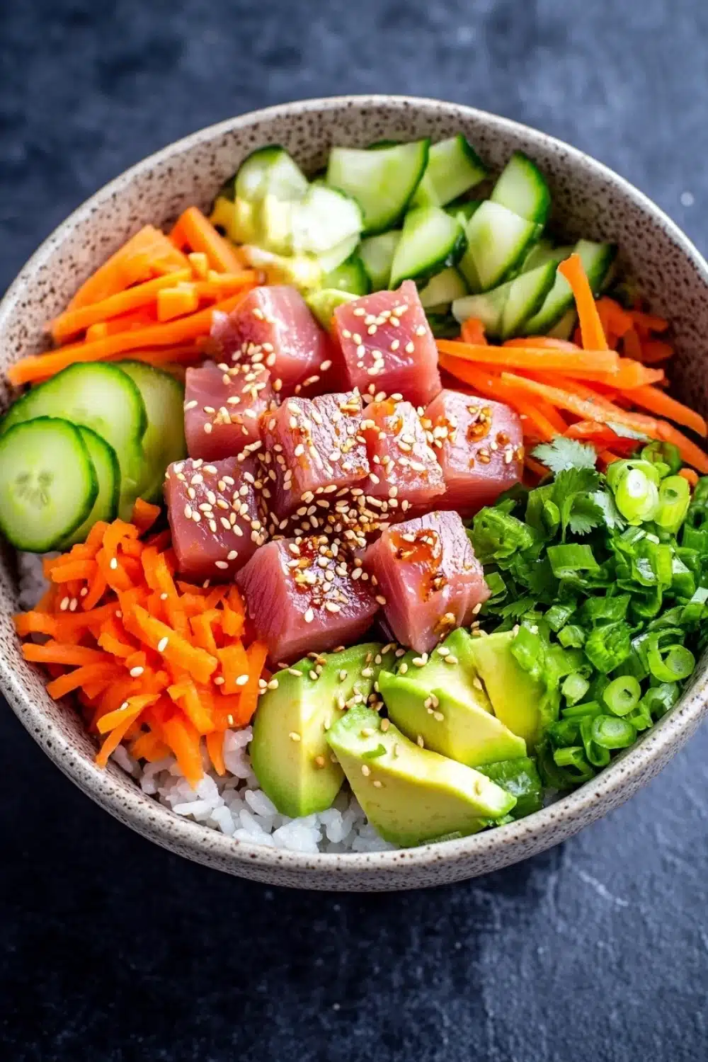 Tuna poke bowl spicy - the image is a close-up of a bowl of food. the bowl is made of a light-colored ceramic material and is placed on a dark grey countertop. inside the bowl, there are various ingredients such as sliced cucumbers, sliced carrots, sliced avocado, and chopped green onions. on top of the vegetables, there is a layer of sesame seeds. the ingredients are arranged in a colorful and appetizing manner.