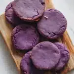 Ube shortbread cookie - the image shows a wooden cutting board with six round, purple cookies on it. the cookies appear to be freshly baked and have a crumbly texture. they are arranged in a scattered manner on the board, with some overlapping each other. the cutting board is placed on a white marble countertop, and there is a green leaf on the right side of the image. the background is blurred, but it appears to be a kitchen countertop.