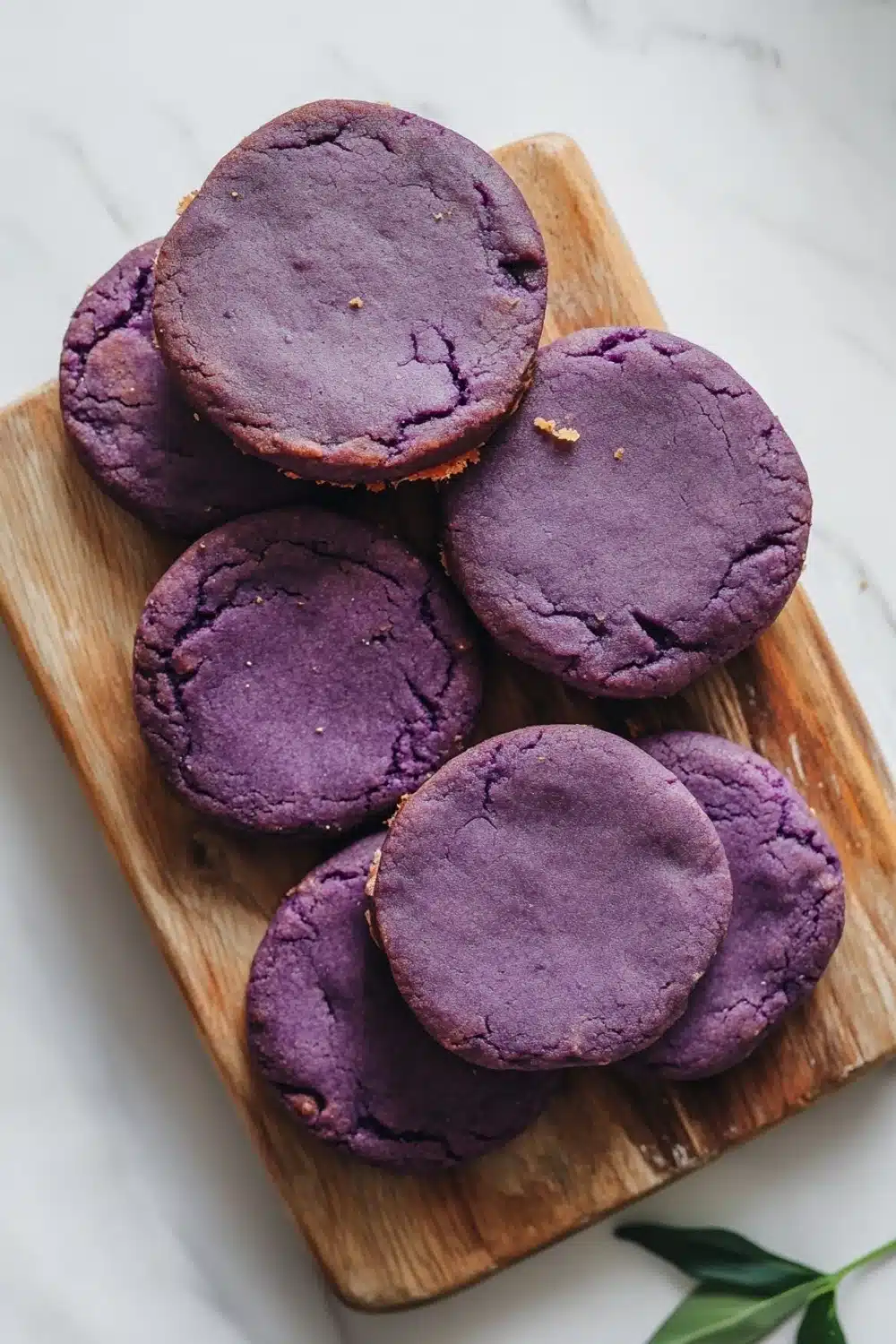 Ube shortbread cookie - the image shows a wooden cutting board with six round, purple cookies on it. the cookies appear to be freshly baked and have a crumbly texture. they are arranged in a scattered manner on the board, with some overlapping each other. the cutting board is placed on a white marble countertop, and there is a green leaf on the right side of the image. the background is blurred, but it appears to be a kitchen countertop.