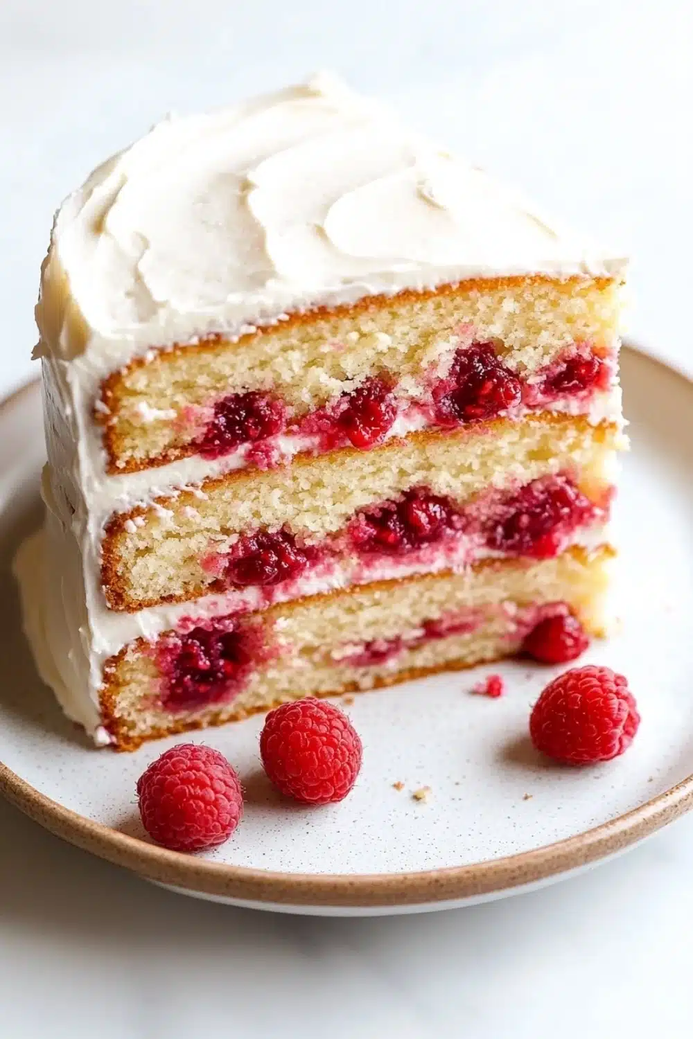 Vanilla cake with raspberry filling - the image is of a slice of cake on a white plate. the cake has a golden brown crust and is topped with a generous layer of white frosting. the frosting is swirled in a circular pattern and is drizzled over the top of the cake. there are also a few fresh raspberries scattered around the plate, adding a pop of color to the dish. the background is plain white, making the cake stand out.
