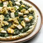 Vegan spinach artichoke dip with tofu - the image is a close-up of a pizza on a white marble countertop. the pizza is round and has a golden-brown crust. on top of the pizza, there are several pieces of artichokes arranged in a circular pattern. the artichoke pieces are golden brown and appear to be seasoned with herbs and spices. there are also small pieces of feta cheese scattered throughout the pizza. the overall appearance is fresh and appetizing.