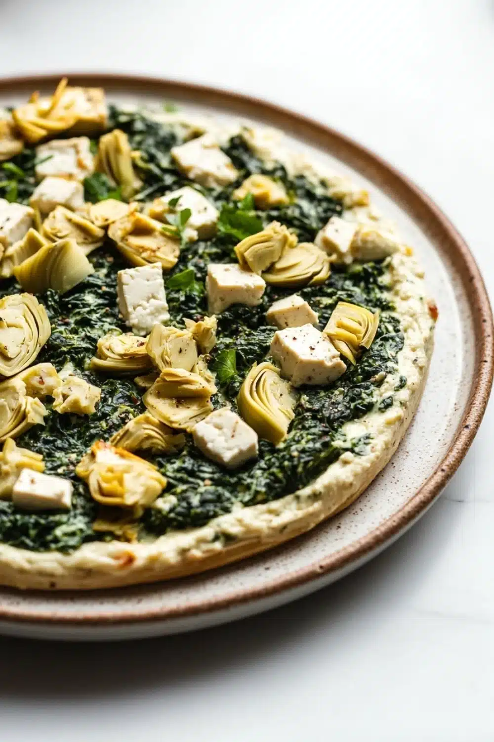 Vegan spinach artichoke dip with tofu - the image is a close-up of a pizza on a white marble countertop. the pizza is round and has a golden-brown crust. on top of the pizza, there are several pieces of artichokes arranged in a circular pattern. the artichoke pieces are golden brown and appear to be seasoned with herbs and spices. there are also small pieces of feta cheese scattered throughout the pizza. the overall appearance is fresh and appetizing.