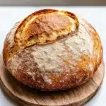 Vegan yeast bread - the image shows a round loaf of bread on a wooden cutting board. the bread appears to be freshly baked, with a golden brown crust and a soft, fluffy interior. the loaf is placed on a white marble countertop, and the cutting board is placed in front of it. the background is blurred, making the bread the focal point of the image.