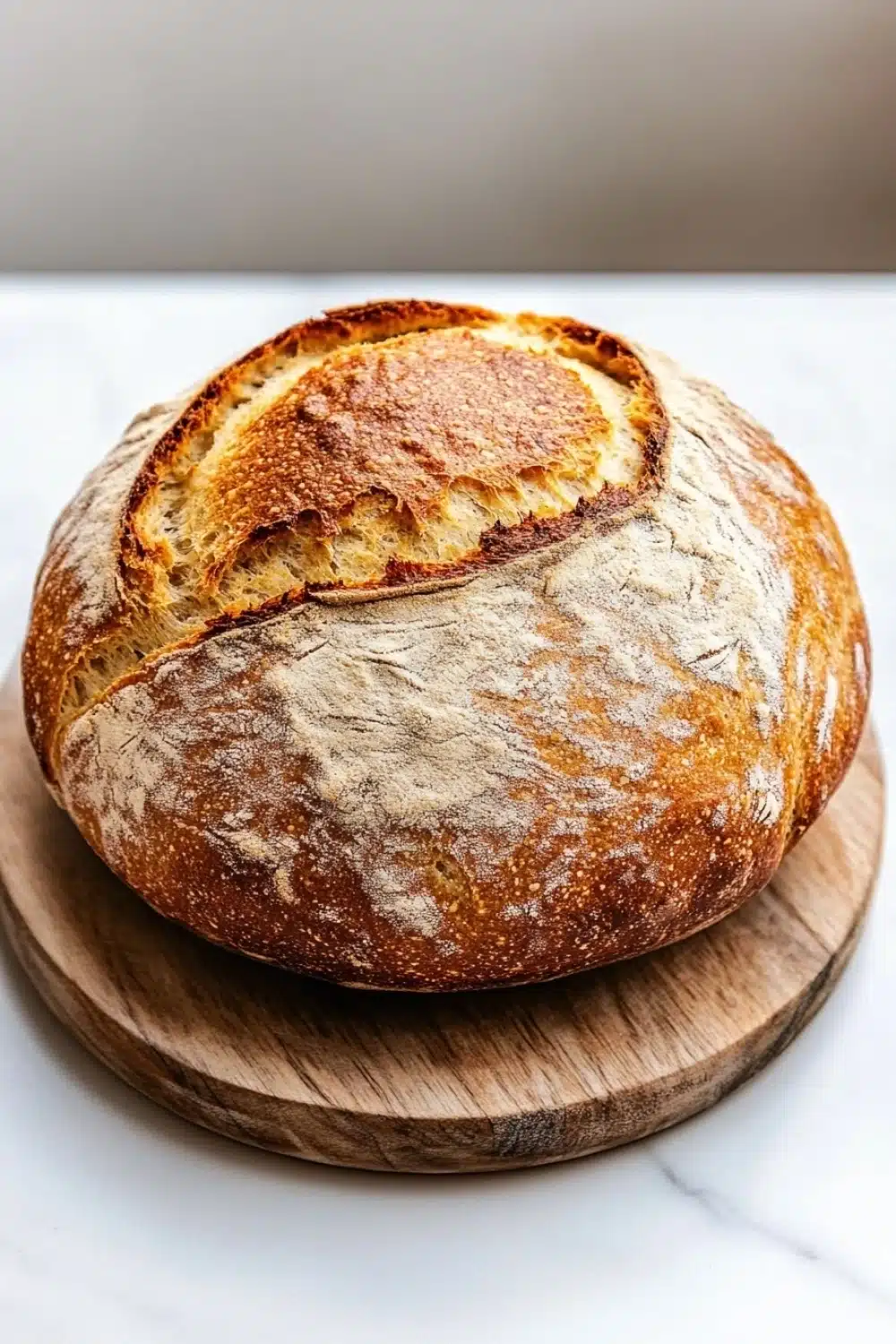 Vegan yeast bread - the image shows a round loaf of bread on a wooden cutting board. the bread appears to be freshly baked, with a golden brown crust and a soft, fluffy interior. the loaf is placed on a white marble countertop, and the cutting board is placed in front of it. the background is blurred, making the bread the focal point of the image.