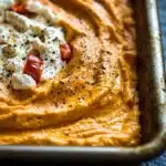 Whipped feta and roasted red pepper dip - the image shows a rectangular baking dish filled with a creamy orange-colored dip. the dip appears to be thick and smooth, with a smooth texture. on top of the dip, there are small pieces of red bell peppers and a dollop of white cream. the dish is sitting on a dark blue surface, possibly a table or countertop. the overall color scheme of the image is warm and inviting.