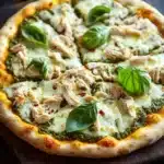 White chicken pesto pizza - the image is of a freshly baked pizza on a wooden cutting board. the pizza has a golden brown crust and is topped with melted white cheese, chunks of chicken, and fresh basil leaves. there are also a few garlic cloves scattered around the cutting board, suggesting that the pizza is ready to be served. the background is dark, making the pizza stand out.