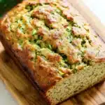 Zucchini bread extra moist - the image shows a freshly baked zucchini bread on a wooden cutting board. the bread is golden brown in color and has a crumbly texture on top. it appears to be freshly baked and has small pieces of zucchinis scattered throughout. on the right side of the cutting board, there is a whole zucchi, which is a bright green color. the cutting board is placed on a white marble countertop.