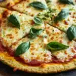 Zucchini crust pizza keto - the image is of a freshly baked pizza on a wooden cutting board. the pizza has a golden brown crust and is topped with melted cheese and fresh basil leaves. the cheese is golden brown and appears to be melted and bubbly. the basil leaves are scattered on top of the cheese, adding a pop of green color to the dish. the background is blurred, but it seems to be a kitchen countertop with a white cloth napkin.