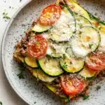 Zucchini tomato ground beef casserole - the image shows a slice of zucchini lasagna on a white plate. the lasagna is topped with sliced zucchinis, sliced tomatoes, minced meat, and a dollop of sour cream. the plate is garnished with fresh herbs and there are two whole tomatoes on the side. the background is a white wooden table.