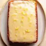 9 x 13 lemon cake - the image shows a rectangular piece of cake on a white plate. the cake appears to be freshly baked and has a golden brown crust on top. the top of the cake is covered in a thick layer of white frosting, which is drizzled with lemon zest. the frosting is spread evenly over the cake, creating a smooth and creamy texture. the plate is round and has small speckles around the edges. the background is a light beige color, making the cake stand out.