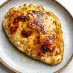 Air fryer chicken breast honey mustard - the image shows a plate with a piece of grilled chicken on it. the chicken is golden brown and appears to be seasoned with herbs and spices. the plate is light blue with a speckled pattern around the edges. the background is a white marble countertop.
