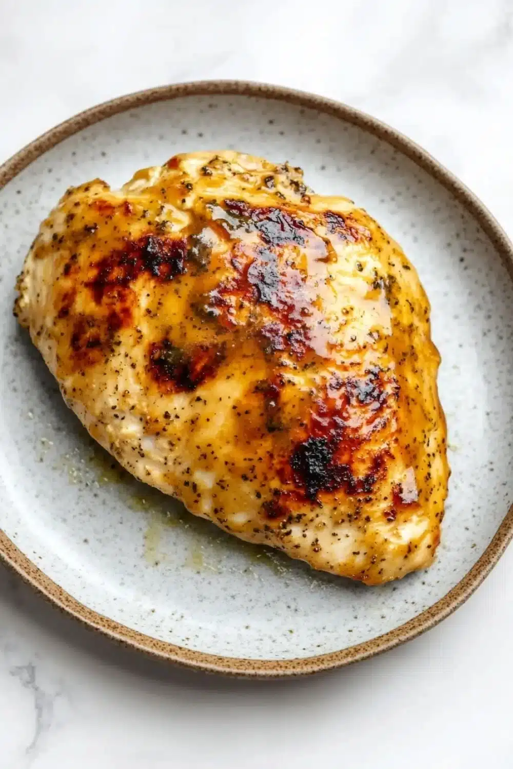 Air fryer chicken breast honey mustard - the image shows a plate with a piece of grilled chicken on it. the chicken is golden brown and appears to be seasoned with herbs and spices. the plate is light blue with a speckled pattern around the edges. the background is a white marble countertop.