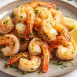 Air fryer peel and eat shrimp - the image shows a plate of cooked shrimp on a white tablecloth. the shrimp are orange in color and appear to be seasoned with herbs and spices. they are arranged in a circular pattern on the plate, with some overlapping each other. there are two lemon wedges on the side of the plate and a few sprigs of parsley scattered around the plate. the background is blurred, but it appears to be a kitchen countertop.