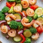 Air fryer shrimp and vegetables - the image is a close-up of a plate of food. the plate is white with a speckled pattern and is placed on a wooden table. the food appears to be a stir-fry dish with shrimp, broccoli, zucchini, red bell peppers, and sliced red onions. the shrimp are arranged in a circular pattern on top of the vegetables, with some overlapping each other. the vegetables are bright green and look fresh and vibrant. the overall appearance of the dish is colorful and appetizing.