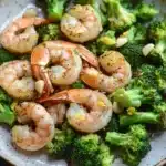 Air fryer shrimp broccoli - the image is a close-up of a plate of cooked shrimp and broccoli. the shrimp are pink and appear to be seasoned with herbs and spices. the broccoli is bright green and looks fresh and healthy. the plate is white with a speckled pattern around the edges. the background is dark, making the colors of the dish stand out.