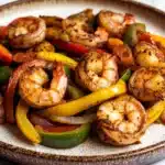 Air fryer shrimp fajitas - the image is a close-up of a plate of grilled shrimp and vegetables. the shrimp are cooked to a golden brown color and appear to be seasoned with herbs and spices. the vegetables are mixed in with the shrimp, including red, yellow, and green bell peppers. the plate is white with a brown rim and is sitting on a white marble countertop. the overall appearance of the dish is colorful and appetizing.