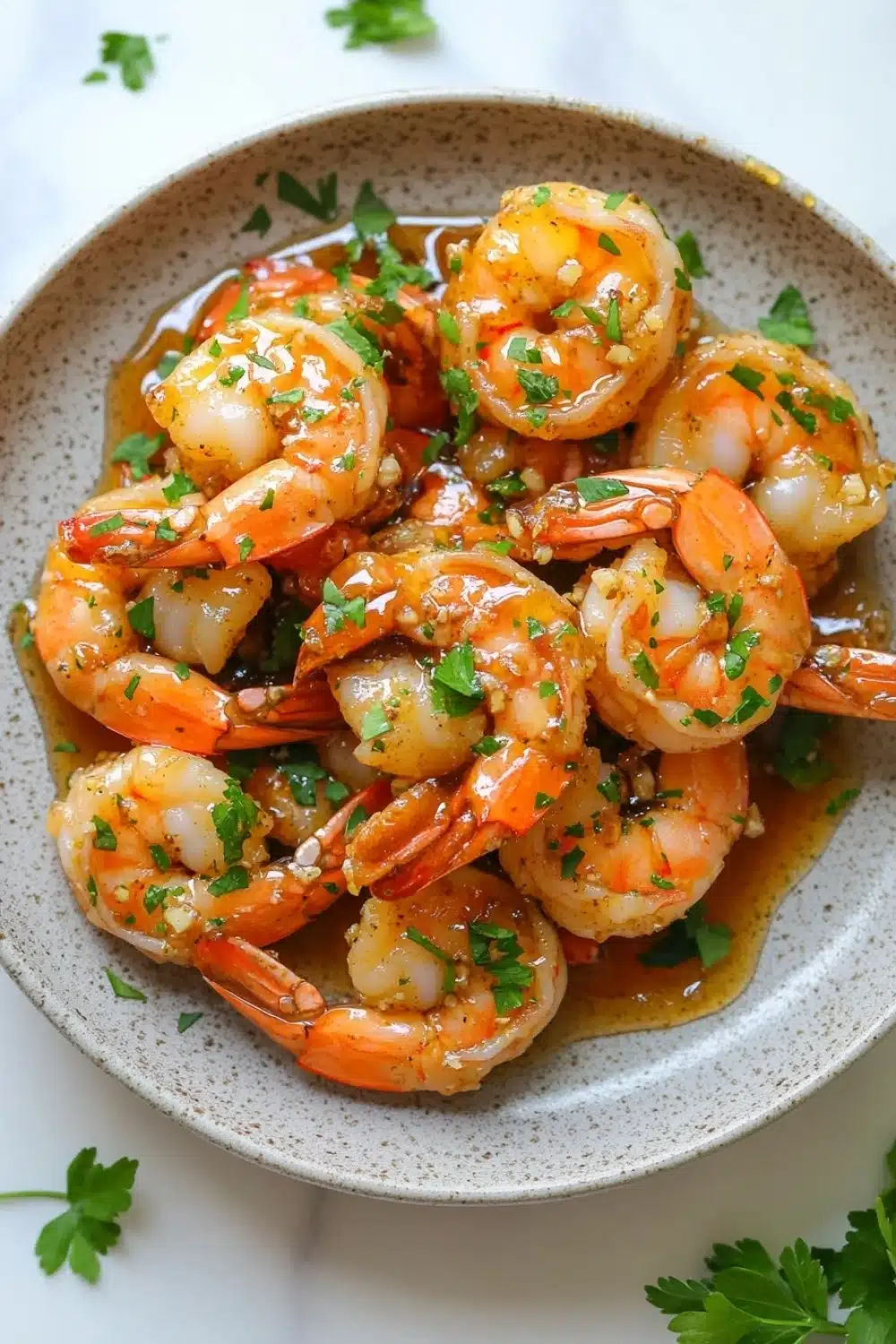 Air fryer shrimp honey garlic - the image shows a plate of cooked shrimp in a creamy sauce. the shrimp are orange in color and appear to be seasoned with herbs and spices. the sauce is a light brown color and is drizzled over the shrimp. the plate is white with a speckled pattern and is garnished with fresh parsley leaves. the background is a white marble countertop.