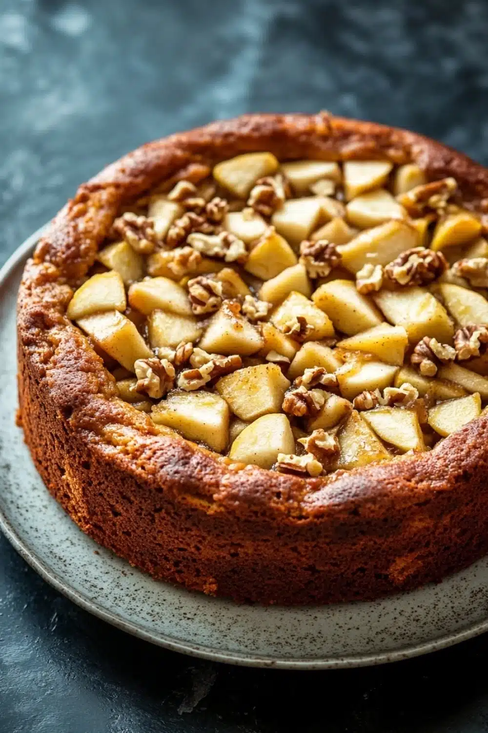 Apple cake from mix boxes - the image is of a round cake on a white plate. the cake appears to be freshly baked and has a golden brown crust on top. on top of the cake, there are sliced apples arranged in a circular pattern. the apples are cut into small cubes and are scattered randomly across the cake. the plate is sitting on a dark grey countertop. the background is blurred, making the cake the focal point of the image.