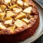 Apple cake gluten free - the image is of a round cake on a white plate. the cake appears to be freshly baked and has a golden brown crust on top. on top of the cake, there are sliced apples arranged in a circular pattern. the apples are cut into small cubes and are covered in a layer of cinnamon sugar. there are also some pecans scattered throughout the cake. the plate is sitting on a dark grey countertop.