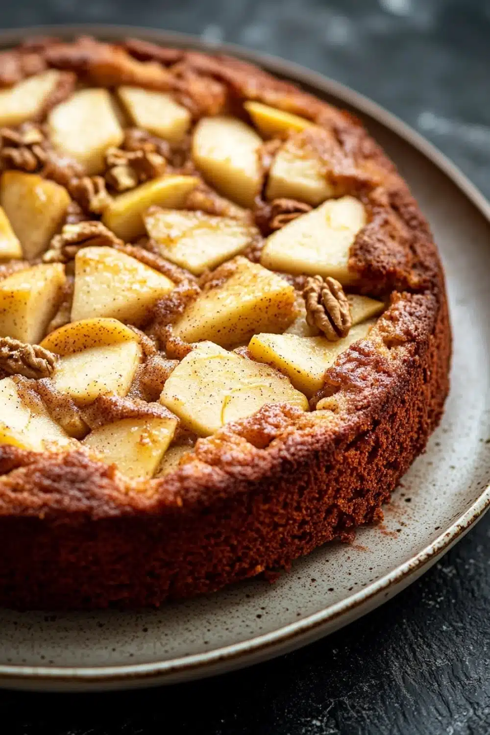 Apple cake gluten free - the image is of a round cake on a white plate. the cake appears to be freshly baked and has a golden brown crust on top. on top of the cake, there are sliced apples arranged in a circular pattern. the apples are cut into small cubes and are covered in a layer of cinnamon sugar. there are also some pecans scattered throughout the cake. the plate is sitting on a dark grey countertop.