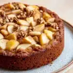 Apple cake no milk - the image is of a round cake on a blue and white plate. the cake appears to be a chocolate cake with a golden brown crust and a layer of caramelized apples on top. the apples are arranged in a circular pattern on the top of the cake, with some of them overlapping each other. there are also some chopped nuts scattered throughout the cake. the plate is sitting on a wooden table.