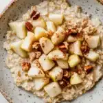 Apple cake oats - the image is a close-up of a bowl of oatmeal with sliced apples and pecans on top. the bowl is made of ceramic and has a light brown color with a speckled pattern. the oatmeal is a creamy white color and appears to be thick and fluffy. the apples are cut into small cubes and are arranged in a circular pattern on top of the oatmeal. there are also some chopped nuts scattered throughout the dish, adding a crunchy texture. the background is a white marble countertop.