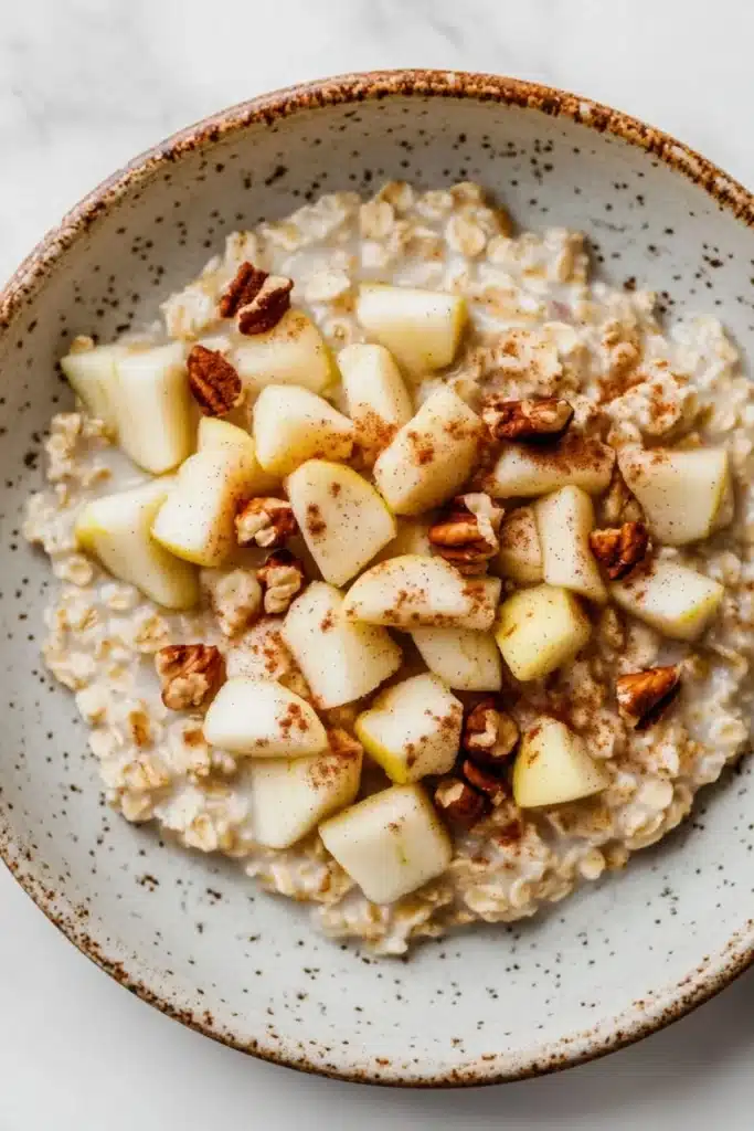 Apple cake oats - the image is a close-up of a bowl of oatmeal with sliced apples and pecans on top. the bowl is made of ceramic and has a light brown color with a speckled pattern. the oatmeal is a creamy white color and appears to be thick and fluffy. the apples are cut into small cubes and are arranged in a circular pattern on top of the oatmeal. there are also some chopped nuts scattered throughout the dish, adding a crunchy texture. the background is a white marble countertop.