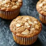 Apple muffin with almond flour - the image shows a group of freshly baked muffins on a black slate surface. the muffins are golden brown in color and have a crumbly texture. they are topped with sliced almonds, giving them a crunchy appearance. the tray they are on is lined with parchment paper, and the background is blurred, making the muffins the focal point of the image.