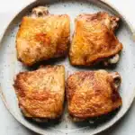 Baked chicken thighs dry rub - the image shows four pieces of grilled chicken on a white marble countertop. the chicken is golden brown and appears to be seasoned with herbs and spices. the pieces are arranged in a circular pattern on the plate, with each piece slightly overlapping the one below it. the plate is round and has a light blue rim. the background is blurred, making the chicken the focal point of the image.