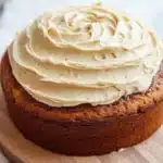 Banana bread birthday cake - the image is of a round cake on a wooden cake stand. the cake appears to be a carrot cake with a golden brown crust. on top of the cake, there is a generous layer of cream cheese frosting that is swirled in a spiral pattern. the frosting is a light beige color and has a smooth texture. the stand is placed on a white marble countertop.