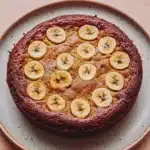 Banana cake allrecipes - the image is of a round cake on a white plate with a light blue rim. the cake appears to be a chocolate cake with a golden brown crust and is topped with sliced bananas arranged in a circular pattern. the bananas are evenly spaced and appear to be freshly baked. the plate is sitting on a wooden table with a beige background.