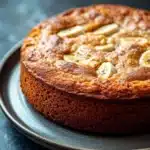 Banana cake no buttermilk - the image is a close-up of a round cake on a gray plate. the cake appears to be freshly baked and has a golden brown crust on top. on top of the cake, there are small pieces of sliced bananas arranged in a circular pattern. the bananas are evenly spaced and appear to be ripe and juicy. the plate is placed on a dark, textured surface, possibly a table or countertop. the background is blurred, making the cake the focal point of the image.