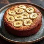 Banana cake snap - the image is of a round cake on a blue plate. the cake appears to be a chocolate cake with a golden brown crust. on top of the cake, there are sliced bananas arranged in a circular pattern. the bananas are yellow and appear to be freshly baked. the plate is sitting on a wooden table with a rustic background. the overall mood of the image is warm and inviting.