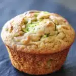 Banana muffin with applesauce and zucchini - the image is a close-up of a freshly baked muffin on a black slate surface. the muffin appears to be golden brown in color and has a crumbly texture. on top of the muffin, there is a layer of green zucchini, which is visible through the crumbs. the zucchinis are arranged in a circular pattern, with some overlapping each other. the background is blurred, making the muffins the focal point of the image.