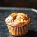 Banana muffin without egg - the image is a close-up of a freshly baked banana muffin on a black baking tray. the muffin is golden brown in color and has a crumbly texture. it is topped with a generous amount of powdered sugar, giving it a shiny appearance. in the center of the muffin, there is a slice of banana that has been cut in half, revealing the soft, fluffy interior. the background is blurred, making the muffins the focal point of the image.