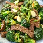 Beef and broccoli stir fry spicy - the image is a close-up of a plate of stir-fry with beef and broccoli. the beef is cooked medium-rare and appears to be seasoned with sesame seeds and green onions. the broccoli is bright green and looks fresh and healthy. the dish is served in a blue and white speckled bowl with a white rim. the background is blurred, making the plate the focal point of the image.