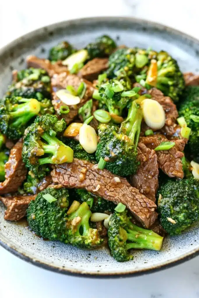 Beef and broccoli stir fry spicy - the image is a close-up of a plate of stir-fry with beef and broccoli. the beef is cooked medium-rare and appears to be seasoned with sesame seeds and green onions. the broccoli is bright green and looks fresh and healthy. the dish is served in a blue and white speckled bowl with a white rim. the background is blurred, making the plate the focal point of the image.
