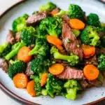 Beef and broccoli stir fry with carrots - the image is a close-up of a plate of stir-fry with beef and broccoli. the beef is cooked medium-rare and appears to be seasoned with herbs and spices. the broccoli is bright green and looks fresh and healthy. there are also small pieces of orange carrots scattered throughout the dish. the plate is white with a brown rim and is sitting on a white tablecloth.