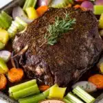 Beef sirloin tip roast slow cooker - the image shows a large piece of cooked meat, possibly a rack of lamb, on a baking tray. the meat appears to be seasoned with herbs and spices, and is garnished with a sprig of rosemary. the tray is filled with various vegetables, including celery, carrots, onions, and potatoes. the vegetables are arranged in a colorful and appetizing manner, with some overlapping each other. the background is a white marble countertop.