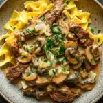 Beef stroganoff crockpot freezer meal - the image is a close-up of a dish in a bowl. the dish appears to be a beef stroganoff, with chunks of beef, mushrooms, and onions mixed in with a creamy sauce. the beef is cooked medium-rare and the mushrooms are sliced and arranged in a circular pattern. the sauce is a light brown color and is drizzled over the top of the beef. the noodles are long and thin, and the dish is garnished with chopped parsley. the bowl is sitting on a dark grey countertop.