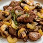 Beef tips freezer meal - the image is a close-up of a plate of food. the plate is white with a speckled pattern and is placed on a wooden table. the food appears to be a dish made with sliced beef, mushrooms, and onions. the beef is cooked medium-rare and has a pinkish-brown color. the mushrooms are sliced into thin slices and are arranged in a pile on top of the onions. there are also small sprigs of fresh herbs scattered throughout the dish. the overall appearance of the dish is savory and appetizing.