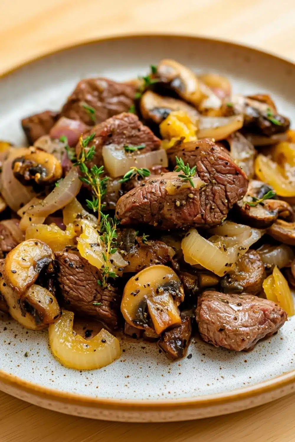 Beef tips freezer meal - the image is a close-up of a plate of food. the plate is white with a speckled pattern and is placed on a wooden table. the food appears to be a dish made with sliced beef, mushrooms, and onions. the beef is cooked medium-rare and has a pinkish-brown color. the mushrooms are sliced into thin slices and are arranged in a pile on top of the onions. there are also small sprigs of fresh herbs scattered throughout the dish. the overall appearance of the dish is savory and appetizing.