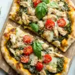 Chicken pesto pizza on naan bread - the image shows two freshly baked pizzas on a wooden cutting board. the pizzas are topped with grilled chicken, cherry tomatoes, and basil leaves. the chicken is cooked to a golden brown color and appears to be seasoned with herbs and spices. the cherry tomatoes are bright red and look ripe and juicy. the basil leaves are scattered around the pizza, adding a pop of green color to the dish. the cutting board is placed on a white marble countertop.