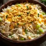 Chicken rice casserole cream soup - the image is a close-up of a bowl of food. the bowl is made of ceramic and is placed on a wooden table. the food appears to be a casserole dish with shredded chicken, cheese, and green onions. the chicken is golden brown and looks crispy on the outside. the cheese is melted and bubbly on top, and there are small pieces of green onions scattered throughout the dish. there are also some sprigs of parsley on the table, adding a pop of green color to the image.