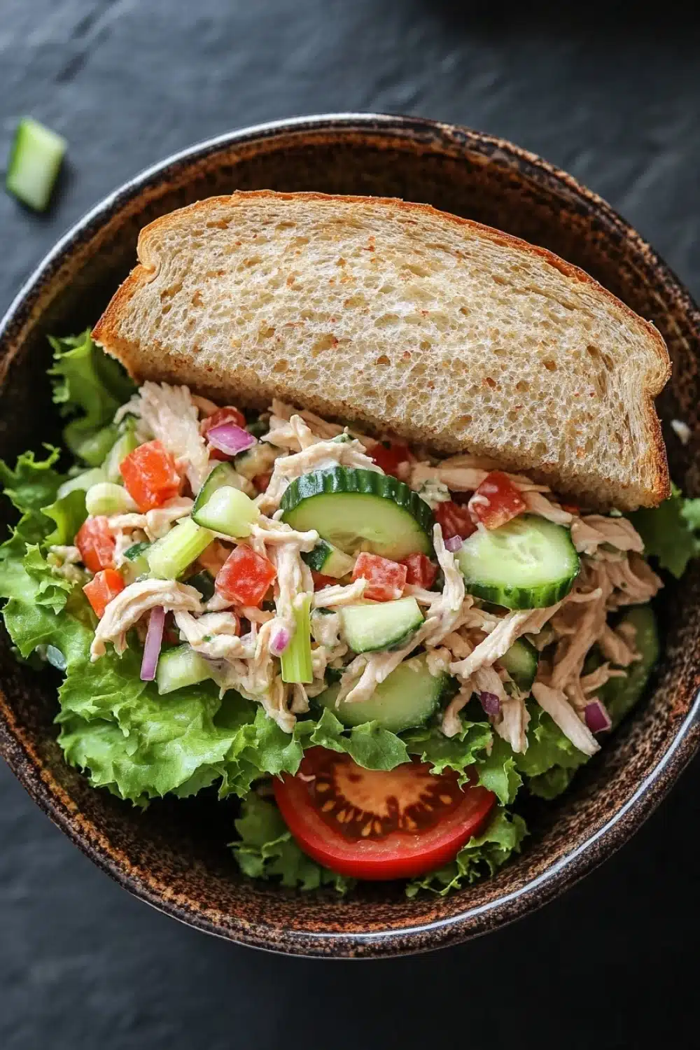 Chicken salad sandwich with veggies - the image shows a bowl of salad with a slice of toasted bread on top. the salad is made up of various types of lettuce, including lettuce, cherry tomatoes, cucumbers, and red onions. the bread is golden brown and appears to be freshly baked. the background is a dark grey countertop. the overall color scheme of the image is warm and inviting.