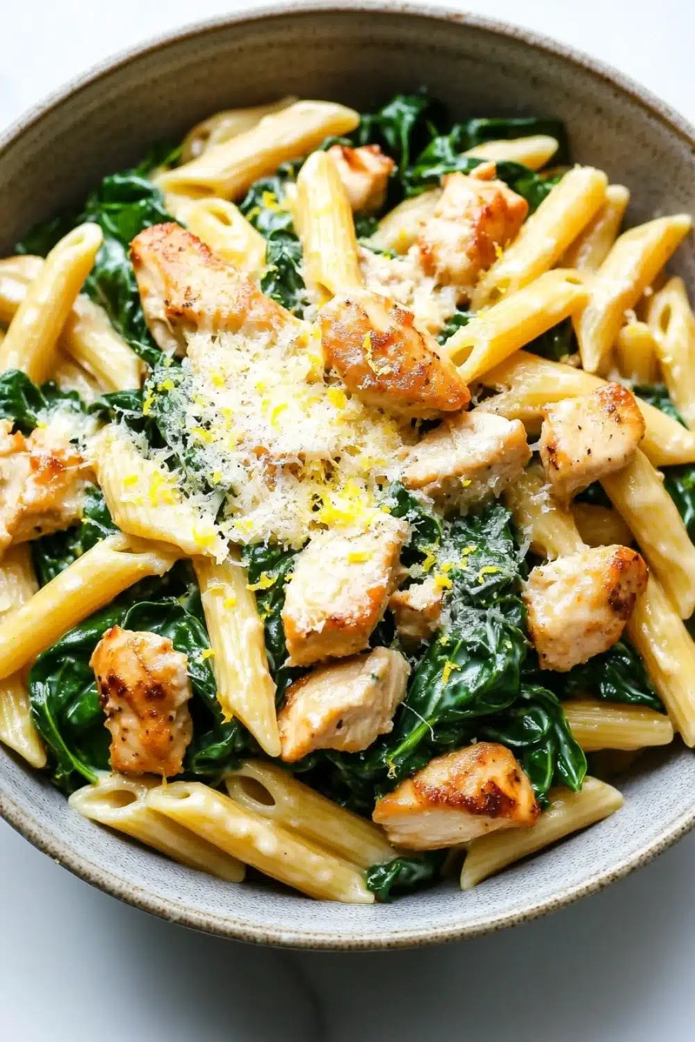 Chicken spinach skillet pasta with lemon parmesan - the image is a close-up of a bowl of pasta dish. the pasta appears to be penne, which is a type of penne pasta, and is cooked until it is golden brown. the dish is garnished with chunks of grilled chicken and fresh spinach. the chicken is cooked to a medium-rare and has a crispy texture. the spinach is a vibrant green color and is scattered throughout the dish, adding a pop of color to the dish. there is also a sprinkle of grated parmesan cheese on top of the chicken, adding texture and flavor. the bowl is made of light-colored ceramic and is sitting on a white marble countertop.