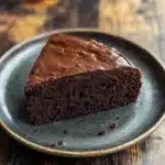 Chocolate banana yoghurt cake - the image shows a slice of chocolate cake on a blue plate. the cake appears to be moist and has a rich, dark chocolate color. it is covered in a thick layer of chocolate frosting, which is spread evenly across the top of the cake. the plate is sitting on a wooden table with a rustic, weathered look. the background is blurred, making the cake the focal point of the image.