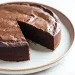 Chocolate cake japanese - the image shows a round chocolate cake on a white plate. the cake has a thick layer of chocolate frosting on top, with a slice taken out of it. the frosting is smooth and shiny, and it appears to be freshly baked. the plate is round and has a light beige rim. the background is a white marble countertop.