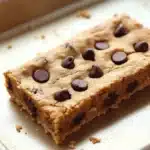Chocolate chip cookie dough bars - the image shows a rectangular piece of chocolate chip cookie bars on a white baking tray. the cookie bars are golden brown in color and have a crumbly texture. they are arranged in a neat row on the tray, with chocolate chips scattered throughout. the tray is sitting on a wooden surface, and there are a few crumbs scattered around the tray.