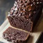 Chocolate zucchini yogurt bread - the image shows a loaf of chocolate bread on a wooden cutting board. the bread is dark brown in color and has a shiny texture. it is topped with small chocolate chips scattered across its surface. a slice of the bread is cut in half, revealing the moist and fluffy interior. the cutting board is placed on a dark wooden table with a rustic background.